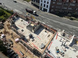 14.08.2025:  Auf der Baustelle sind die Arbeiten am Erdgeschoss in unterschiedlichen Bauabschnitten fortgeschritten.   Im ersten Bauabschnitt wurde die Bodenplatte bereits vollständig hergestellt, und die Betonarbeiten für die tragenden Wände im Erdgeschoss haben begonnen. Erste Wände und Schächte sind betoniert, die Schalungs- und Bewehrungsarbeiten für weitere Wandabschnitte laufen. Das Arbeitsumfeld ist eingerichtet, Material liegt geordnet bereit, und Gerüste an den Außenkanten sichern die laufenden Arbeiten.   Im zweiten Bauabschnitt ist der Rohbau des Erdgeschosses bereits vollständig fertiggestellt. Sämtliche tragenden Wände sind errichtet, und die Baustelle hat nahtlos mit den Rohbauarbeiten im 1. Obergeschoss begonnen. Hier werden aktuell die ersten tragenden Wände des 1. Obergeschosses gestellt, während gleichzeitig vorbereitende Maßnahmen für die das 2.Obergeschoss getroffen werden.