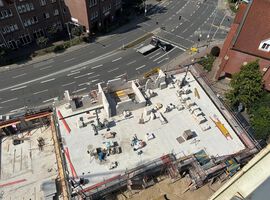 14.08.2025:  Auf der Baustelle sind die Arbeiten am Erdgeschoss in unterschiedlichen Bauabschnitten fortgeschritten.   Im ersten Bauabschnitt wurde die Bodenplatte bereits vollständig hergestellt, und die Betonarbeiten für die tragenden Wände im Erdgeschoss haben begonnen. Erste Wände und Schächte sind betoniert, die Schalungs- und Bewehrungsarbeiten für weitere Wandabschnitte laufen. Das Arbeitsumfeld ist eingerichtet, Material liegt geordnet bereit, und Gerüste an den Außenkanten sichern die laufenden Arbeiten.   Im zweiten Bauabschnitt ist der Rohbau des Erdgeschosses bereits vollständig fertiggestellt. Sämtliche tragenden Wände sind errichtet, und die Baustelle hat nahtlos mit den Rohbauarbeiten im 1. Obergeschoss begonnen. Hier werden aktuell die ersten tragenden Wände des 1. Obergeschosses gestellt, während gleichzeitig vorbereitende Maßnahmen für die das 2.Obergeschoss getroffen werden.