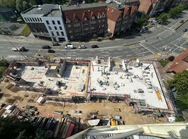 14.08.2025:  Auf der Baustelle sind die Arbeiten am Erdgeschoss in unterschiedlichen Bauabschnitten fortgeschritten.   Im ersten Bauabschnitt wurde die Bodenplatte bereits vollständig hergestellt, und die Betonarbeiten für die tragenden Wände im Erdgeschoss haben begonnen. Erste Wände und Schächte sind betoniert, die Schalungs- und Bewehrungsarbeiten für weitere Wandabschnitte laufen. Das Arbeitsumfeld ist eingerichtet, Material liegt geordnet bereit, und Gerüste an den Außenkanten sichern die laufenden Arbeiten.   Im zweiten Bauabschnitt ist der Rohbau des Erdgeschosses bereits vollständig fertiggestellt. Sämtliche tragenden Wände sind errichtet, und die Baustelle hat nahtlos mit den Rohbauarbeiten im 1. Obergeschoss begonnen. Hier werden aktuell die ersten tragenden Wände des 1. Obergeschosses gestellt, während gleichzeitig vorbereitende Maßnahmen für die das 2.Obergeschoss getroffen werden.